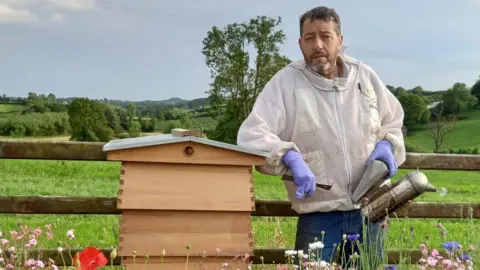 Tom Williamson Tom Williamson wearing a beekeeping jacket with blue jeans standing next to a brown wooden hive. He is holding a silver grey smoker. He has short dark hair and a grey beard.