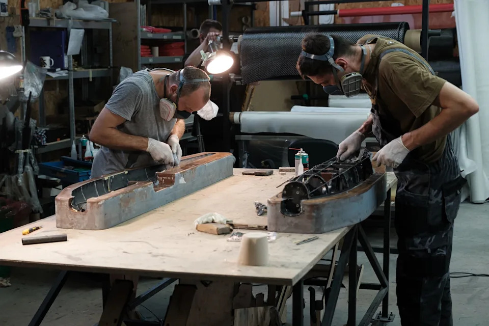 Two men wearing short-sleeved t-shirts and masks bend over a table where they are building objects inside a workroom