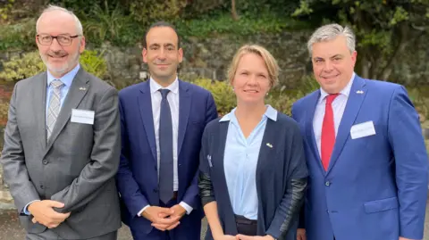 BBC Four people who are officials from France and Guernsey are pictured standing shoulder to shoulder and they are all smiling at the camera. There are three men and one woman. The men are all wearing suits. The woman is wearing a blue shirt with a dark blue cardigan.