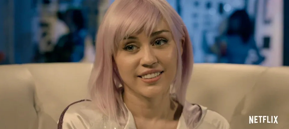 Miley Cyrus with pink hair, sitting on a couch