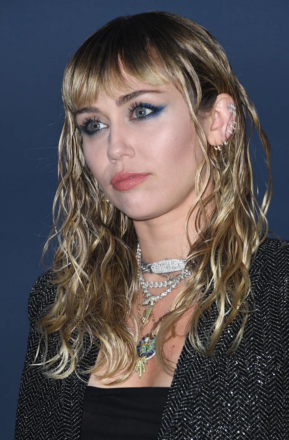 Miley Cyrus is wearing a black jacket with sparkly details, a black top, and a necklace