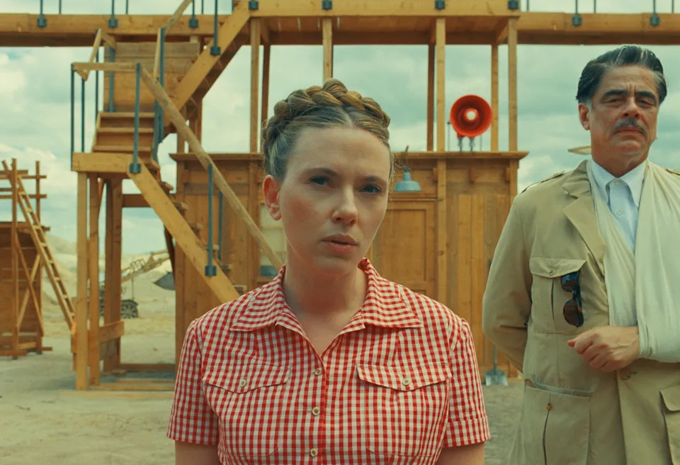Scarlett Johansson is wearing a red plaid shirt. Benicio Del Toro is standing next to her in a trench coat and arm sling. Wooden stairs and buildings are behind them