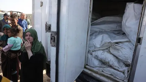 AFP A Palestinian woman stands next to a refrigerated lorry carrying the remains of unidentified Palestinians whose bodies were returned by Israel in exchange for dead Israeli hostages, outside Naser hospital, in Khan Younis, southern Gaza (22 October 2025)