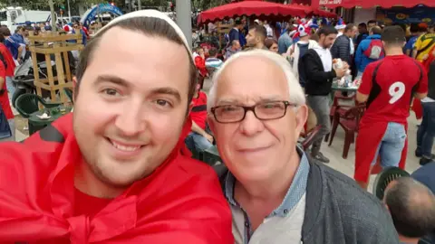 Family handout A man in draped in an Albanian flag, which has a red background and a two-headed black eagle. He is standing next to a man, with white hair, and a grey cardigan and grey shirt. They are attending an event.