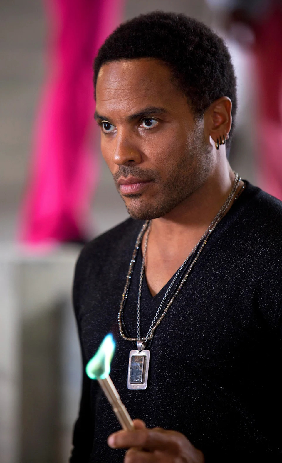 Lenny Kravitz is wearing a black shirt, multiple necklaces, and holding a flaming stick in "The Hunger Games"