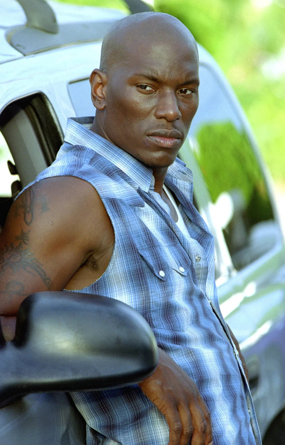Tyrese Gibson is wearing a plaid shirt with no sleeves as he leans against a car