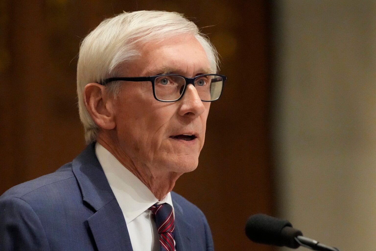 An older man with white hair and glasses, dressed in a blue suit and red striped tie, speaks into a microphone.