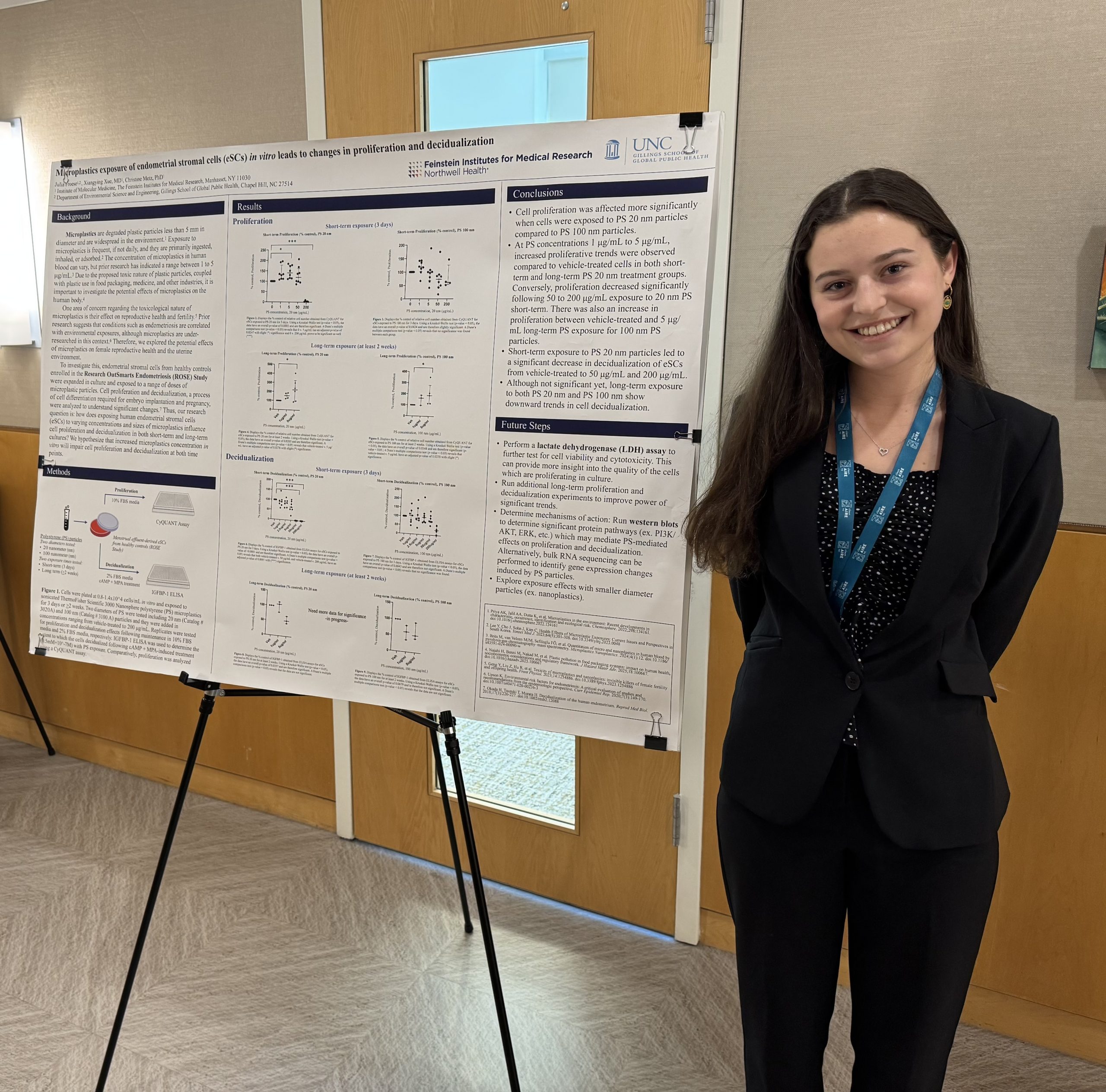 A smiling young woman in a dark pantsuit stands beside a medical research poster.