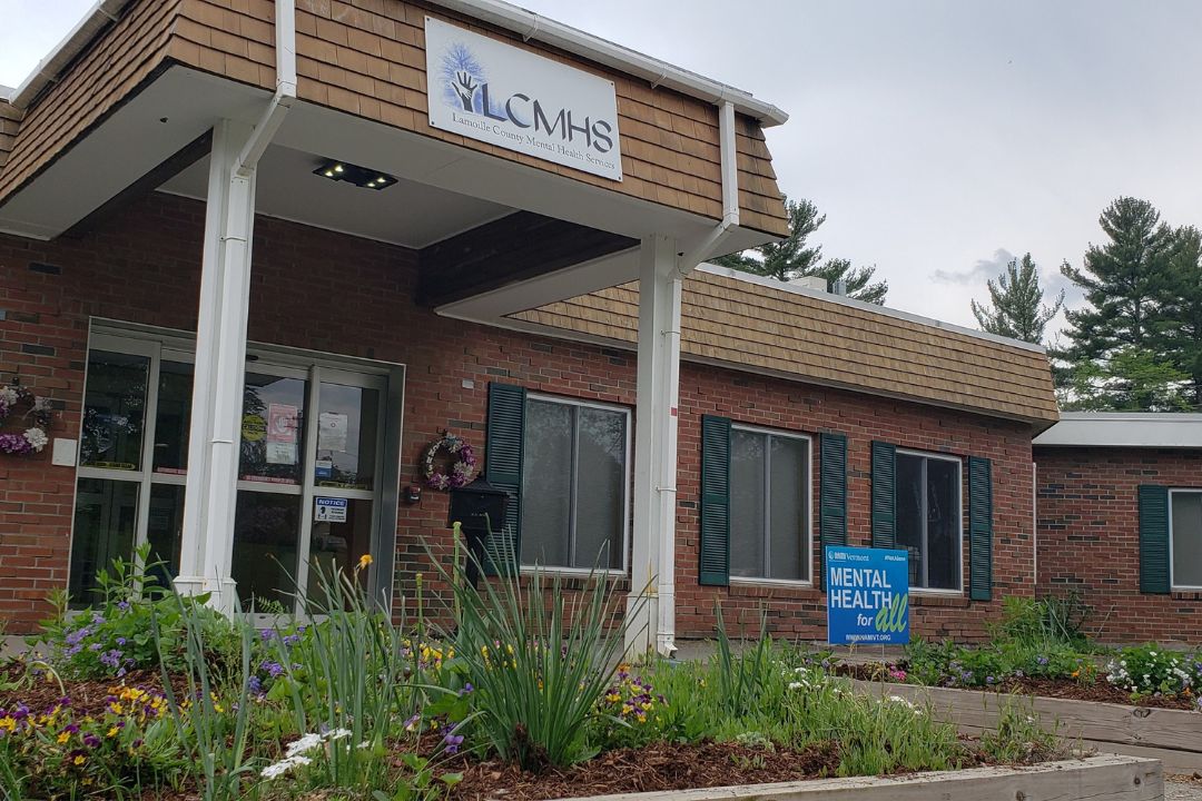 A brick building with a sign reading "LCMHS" above the entrance and a yard sign that says "Mental Health for All" among landscaped plants.