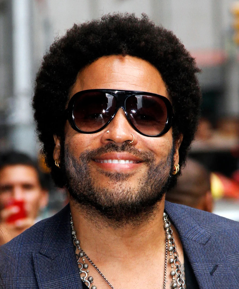 Lenny Kravitz is wearing a blue plaid jacket, multiple necklaces, and is smiling while outside in New York City