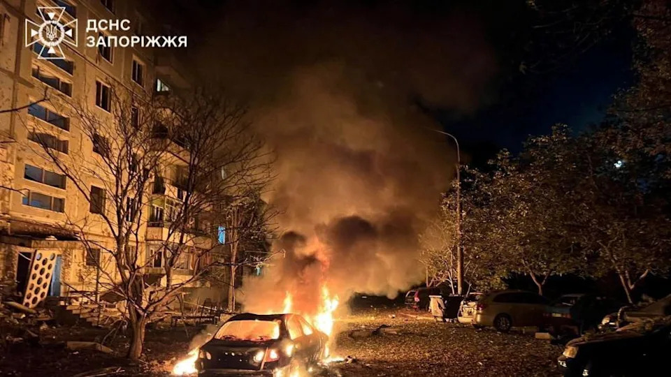 Flames rising from a car on fire in front on an apartment building, during the night.