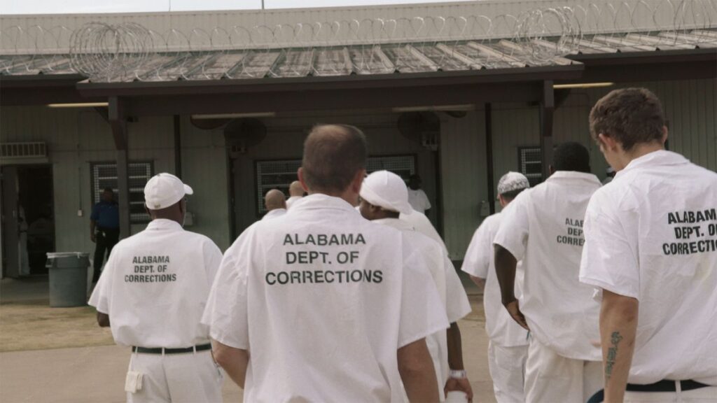 Several men wearing white prison uniforms with the words “Alabama Dept. of Corrections” printed on their backs walk toward a building.