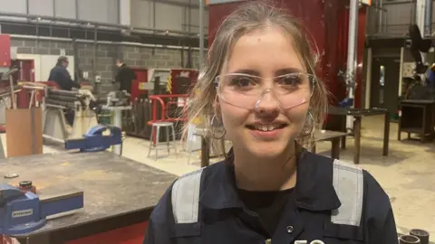 BBC A young woman with long blond hair tied up smiles looking at the camera a she stands in a workshop. She is wearing blue coveralls and safety glasses. Two men are at a work table in the background behind her. 