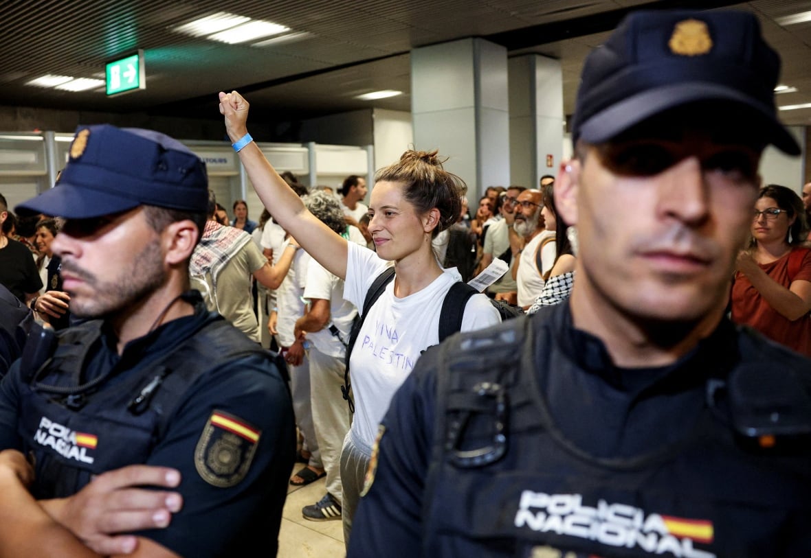 A woman holds up a fist while wearing a white t-shirt with Viva Palestine handwritten on it. As Spanish police hold up a crowd.