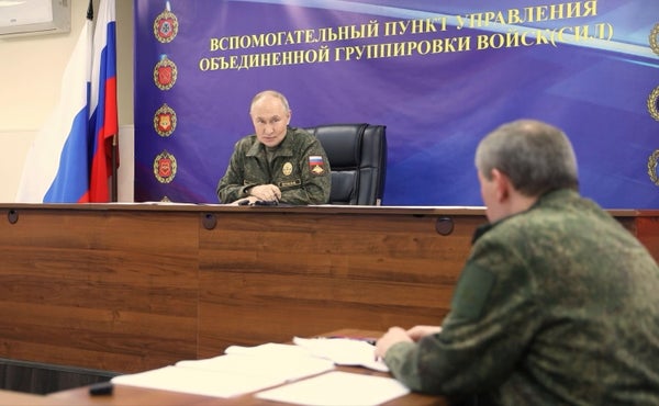 Vladimir Putin sits on a rostrum in fatigues talking to a man in fatigues at a table to his left