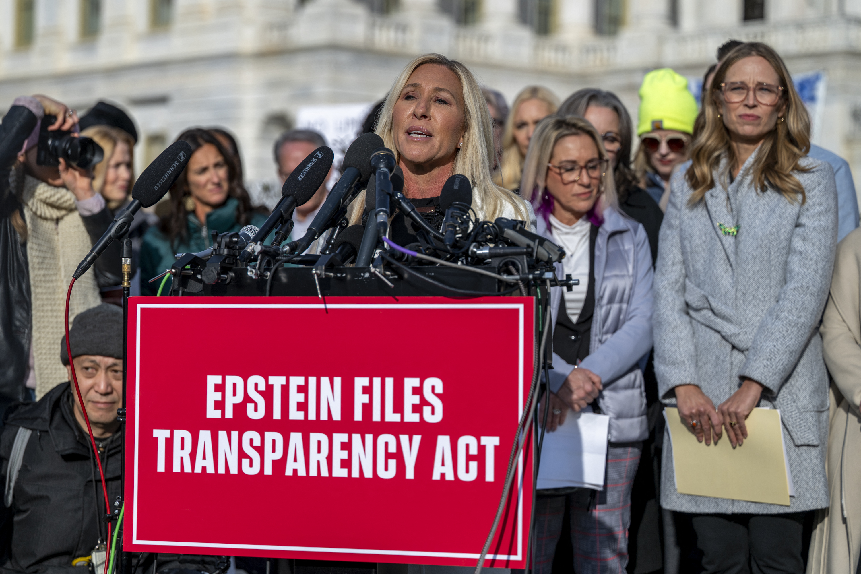 US Representative Marjorie Taylor Greene speaking at a press conference with a red sign in front that reads "Epstein Files Transparency Act."