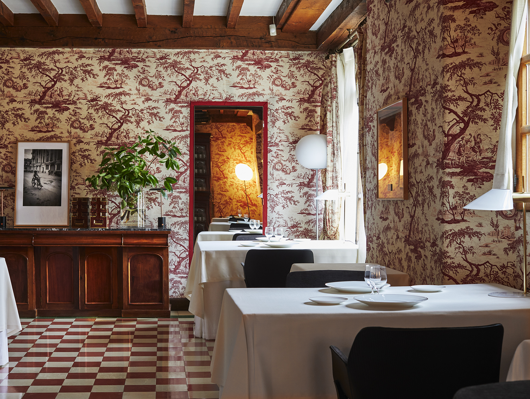 The dining room of Cenador de Amós restaurant with red and white toile wallpaper, white tablecloths, and a red and white checkered floor.