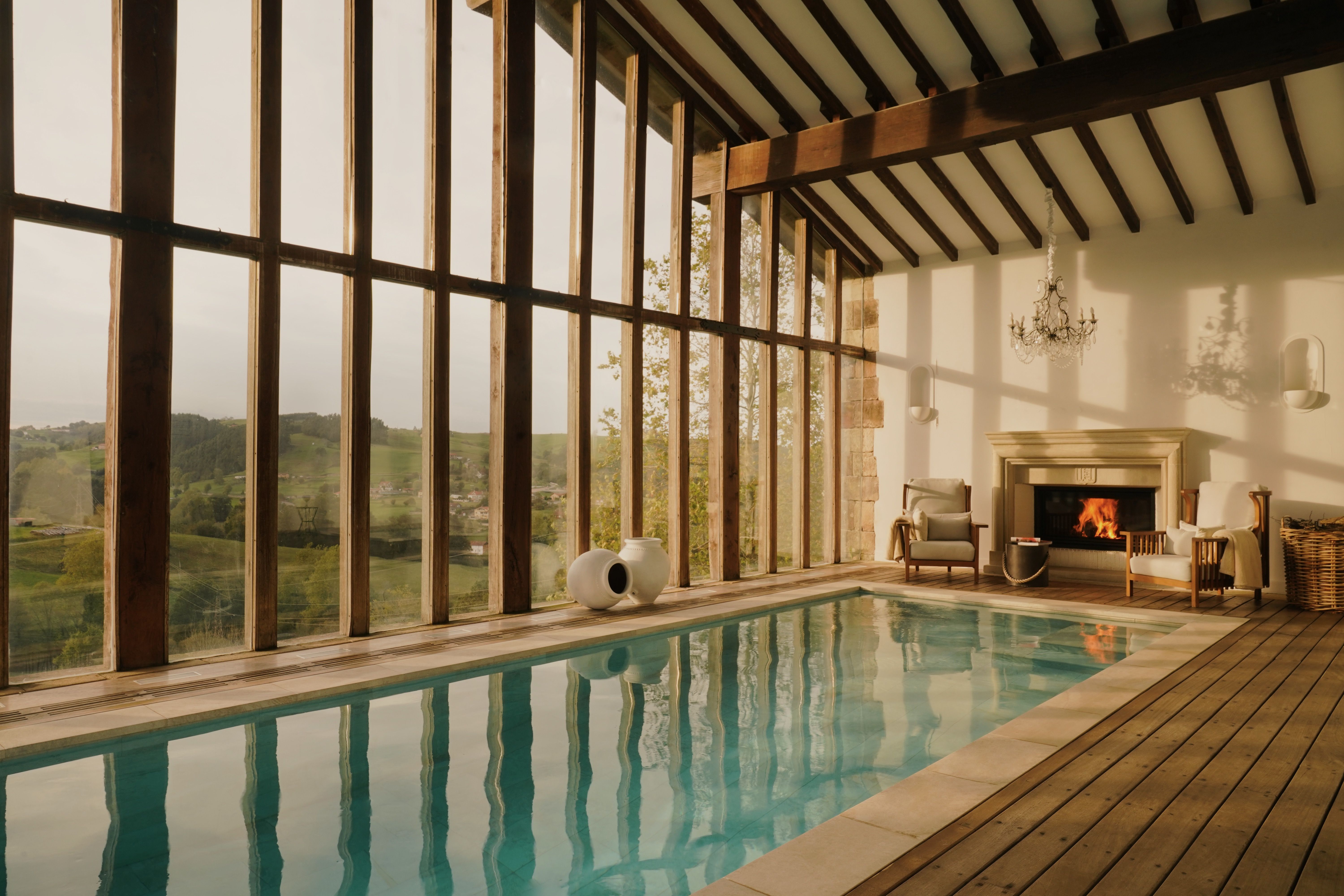 Indoor swimming pool with large windows overlooking a green landscape and a fireplace.