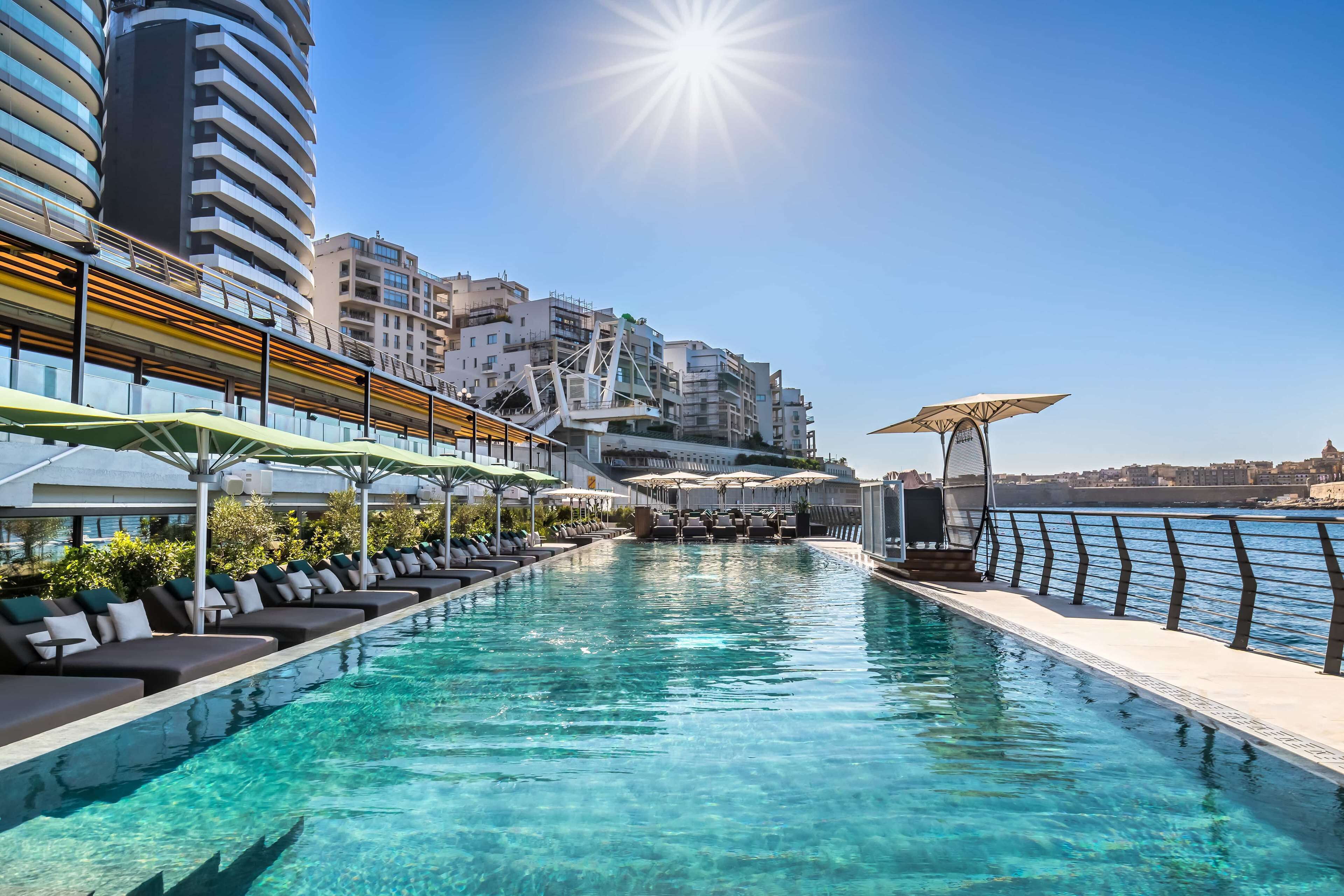 An outdoor pool at Barceló Fortina Malta with lounge chairs and umbrellas, overlooking the sea and a city.