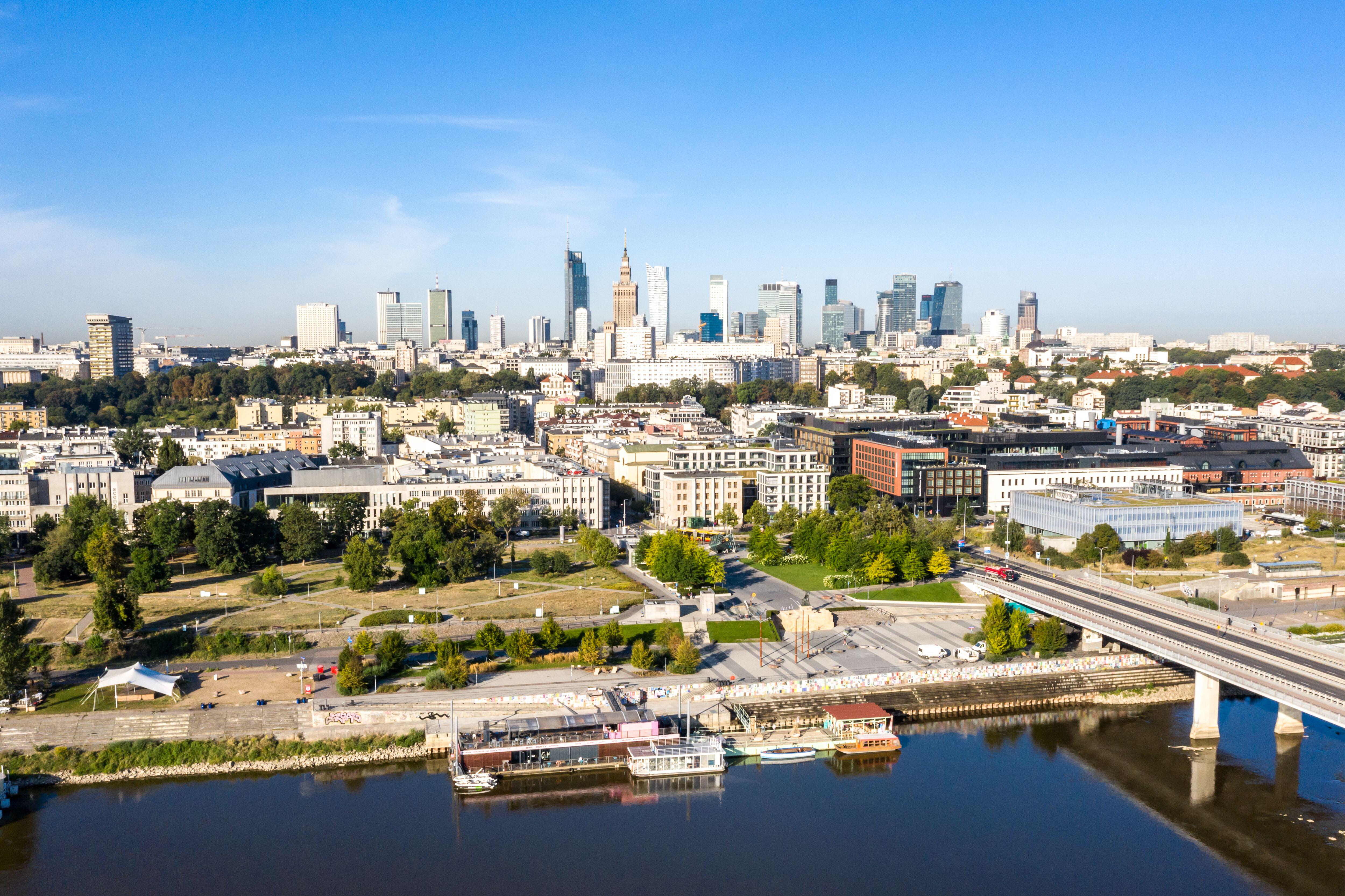Aerial view of Warsaw's financial center, with skyscrapers in the background, a bridge over a river, and waterfront parks and buildings in the foreground.