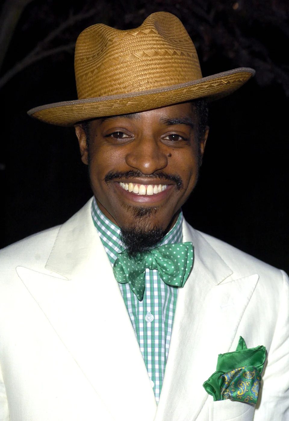 Person in a wide-brimmed straw hat, wearing a white suit, green checkered shirt, green bow tie, and pocket square, smiles at the camera