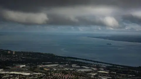 Getty Images A large body of water separates two large plots of land. Their is a grey storm cloud hanging over the blue lough. A ferry can be seen sailing across the lough from right to left of the shot. Their is a collection of factories and houses. 