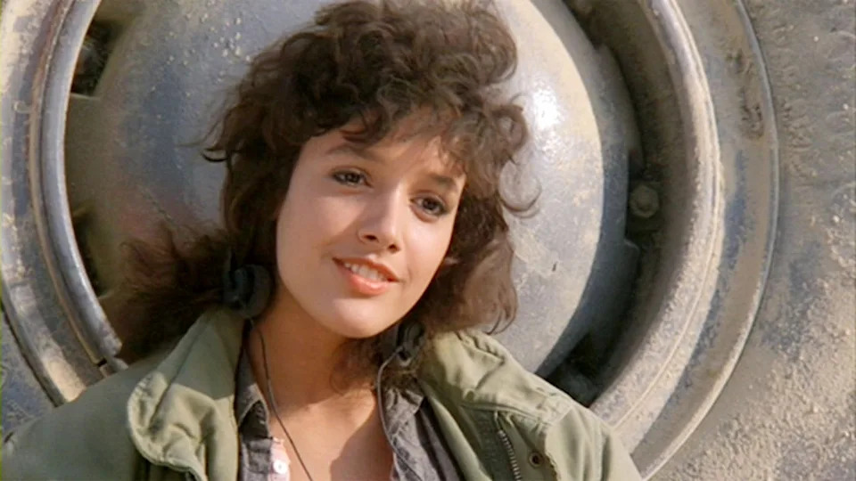 Person with wavy hair smiles, wearing a casual jacket, standing in front of a large industrial object