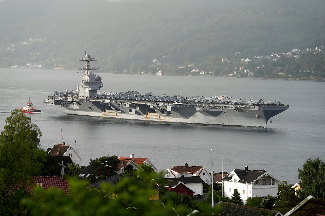 A large military ship is shown in a body of water, with houses seen on a shoreline.