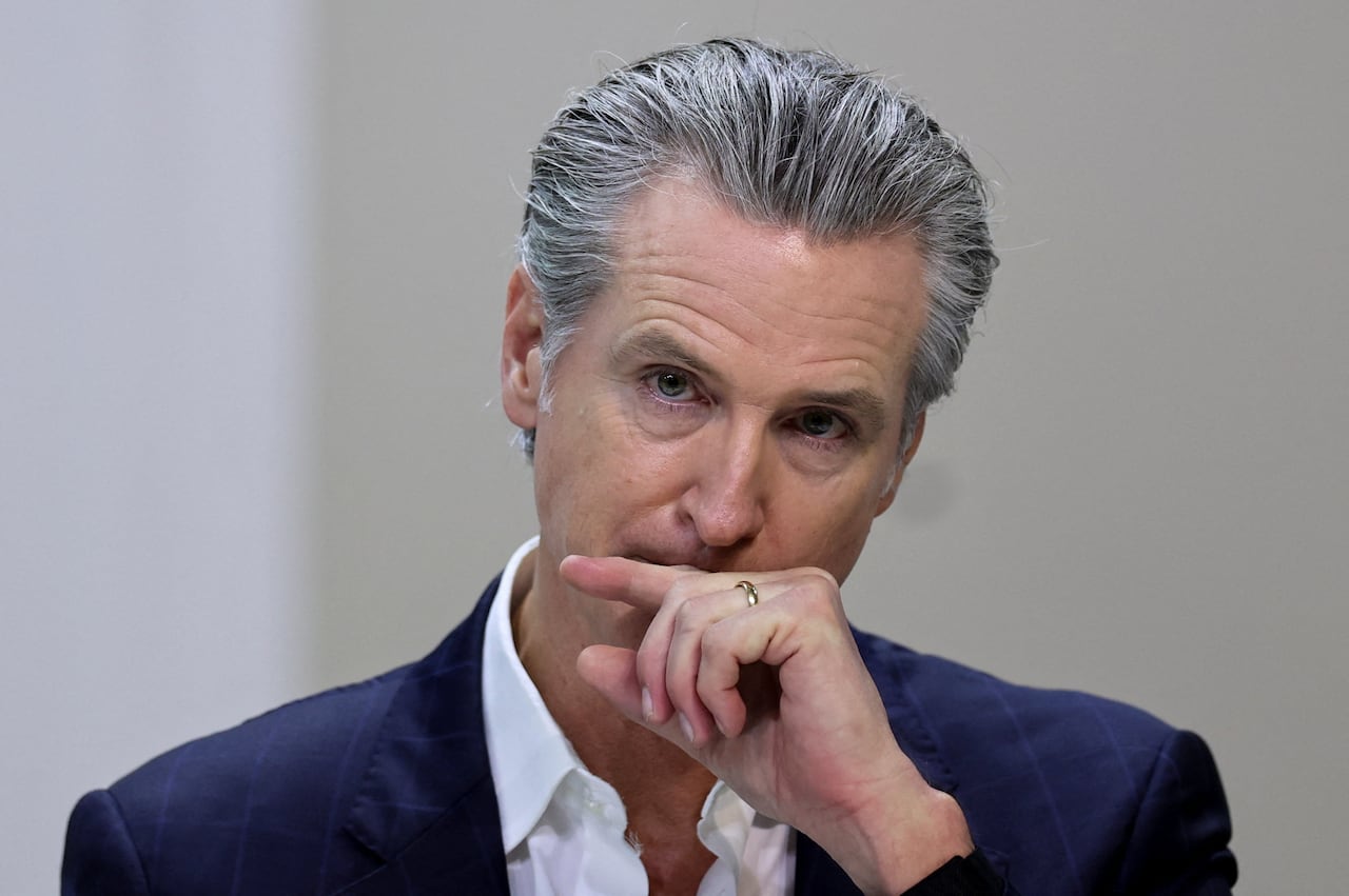 A cleanshaven greyhaired man is shown in closeup with his hand over his chin while wearing a blue blazer. 