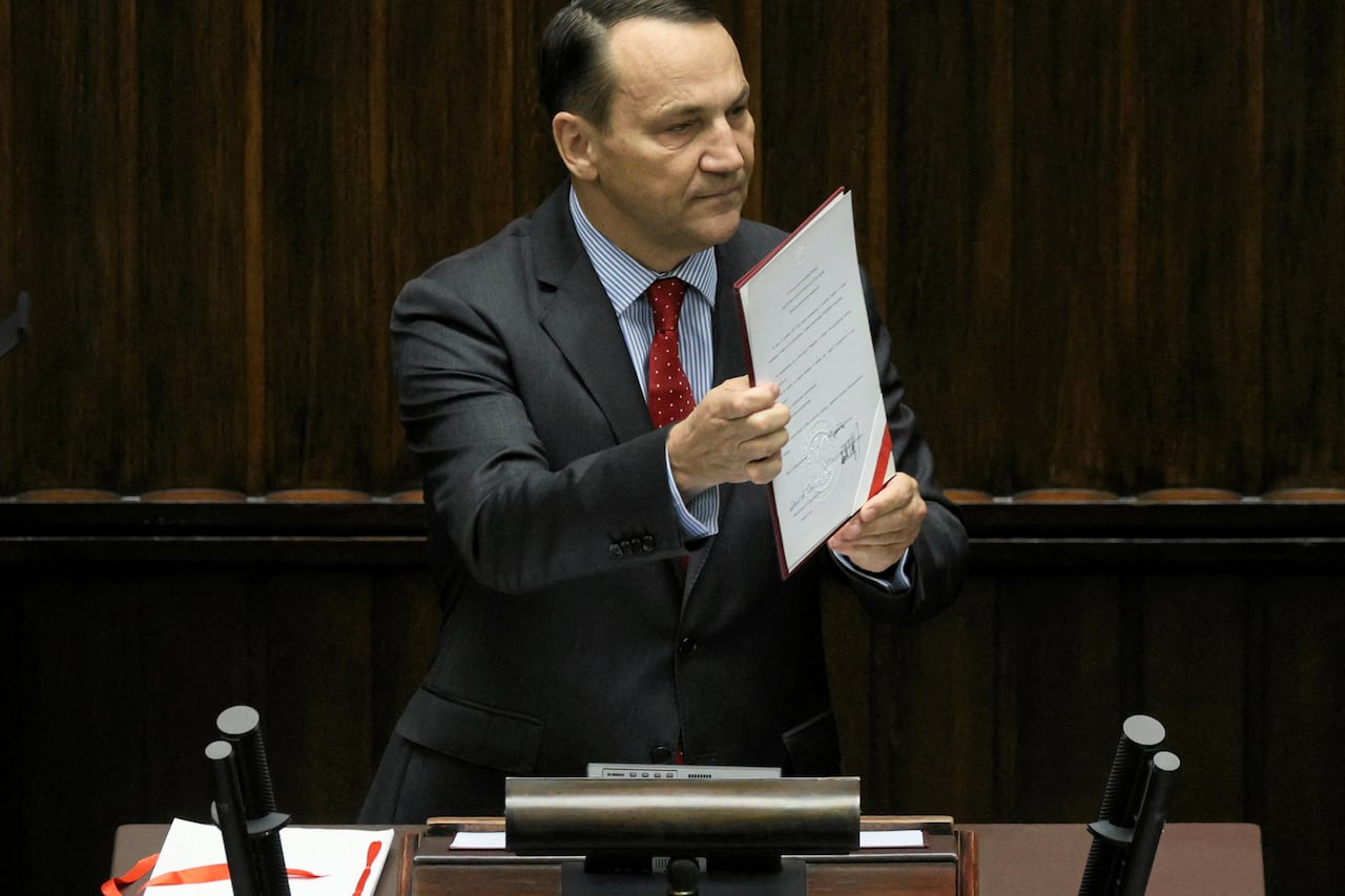 A man in a suit holds up a signed document while standing behind a small desk.