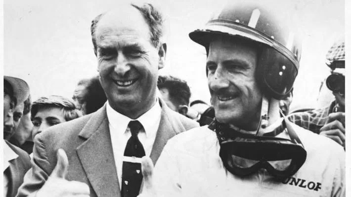 A black and white photo of a man with short hair on the sides wearing a suit and tie and putting his thumb up. Next to him is a younger man wearing a helmet with goggles round his neck looking to the side and smiling. A crowd can be seen behind them.