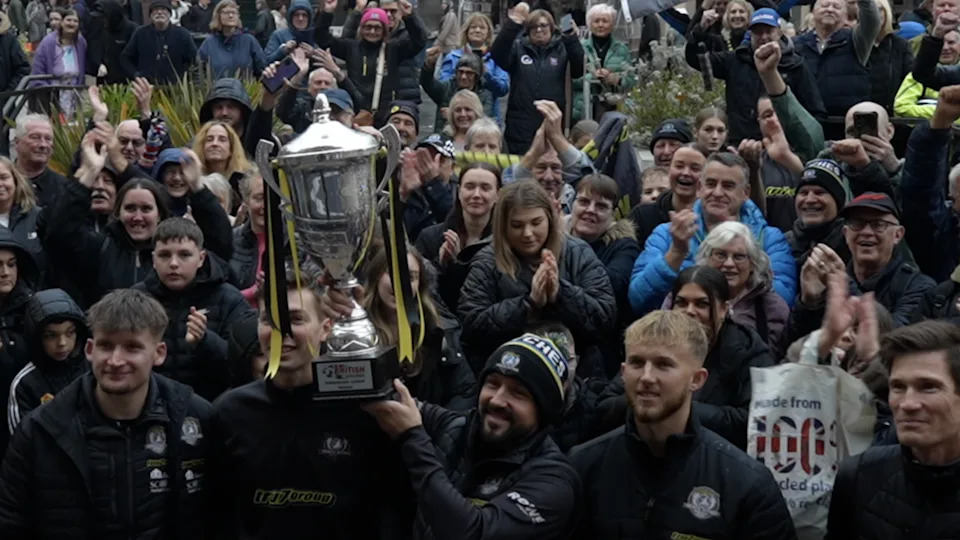 Ipswich Witches team stand in front of a crowd of people with one of the members lifting a large trophy. They are smiling toward the front and some of the fans are clapping and cheering. 