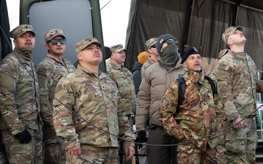 Several soldiers in camouflage uniforms stand in a line looking to the sky.
