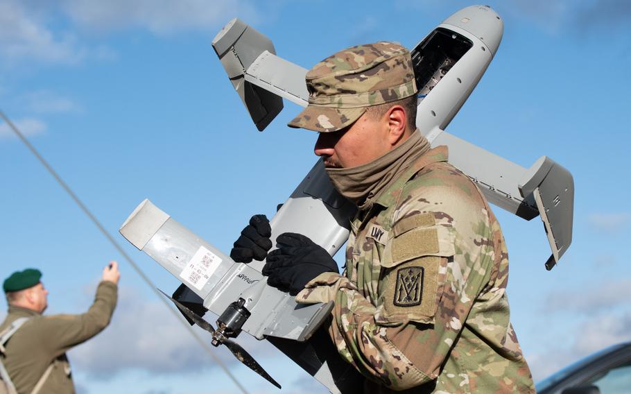 A soldier in camouflage uniform and hat carries a gray-colored military drone on his shoulder.