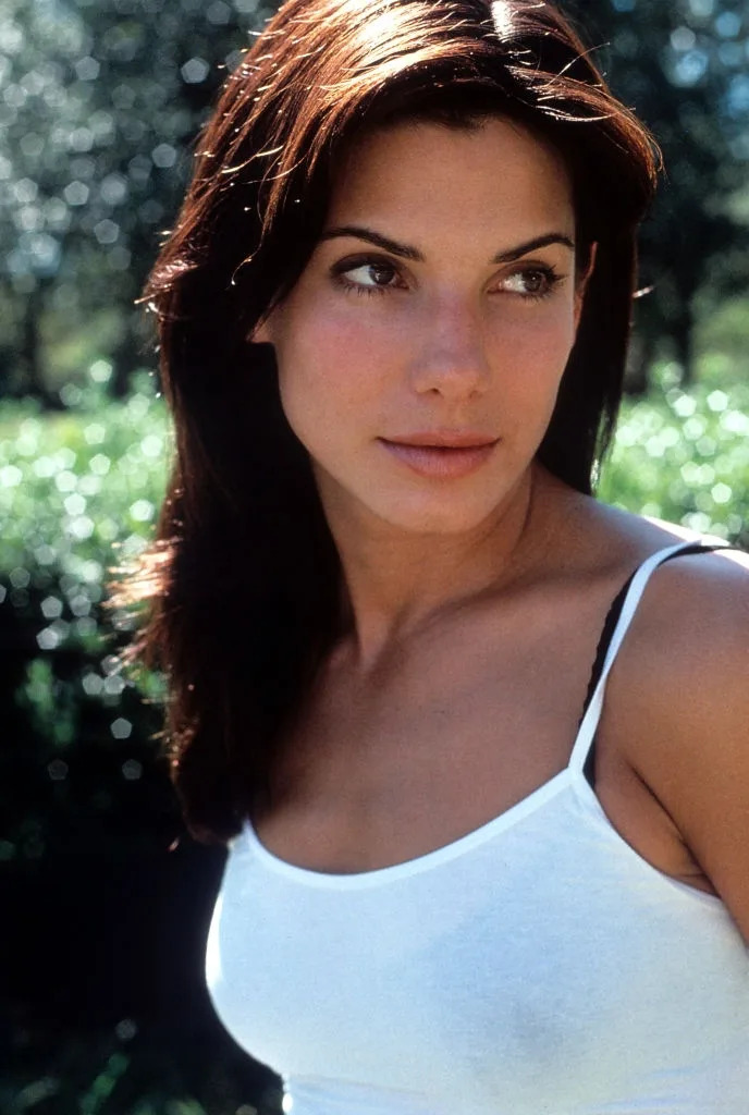 A person with long hair wearing a white tank top outdoors, looking to the side