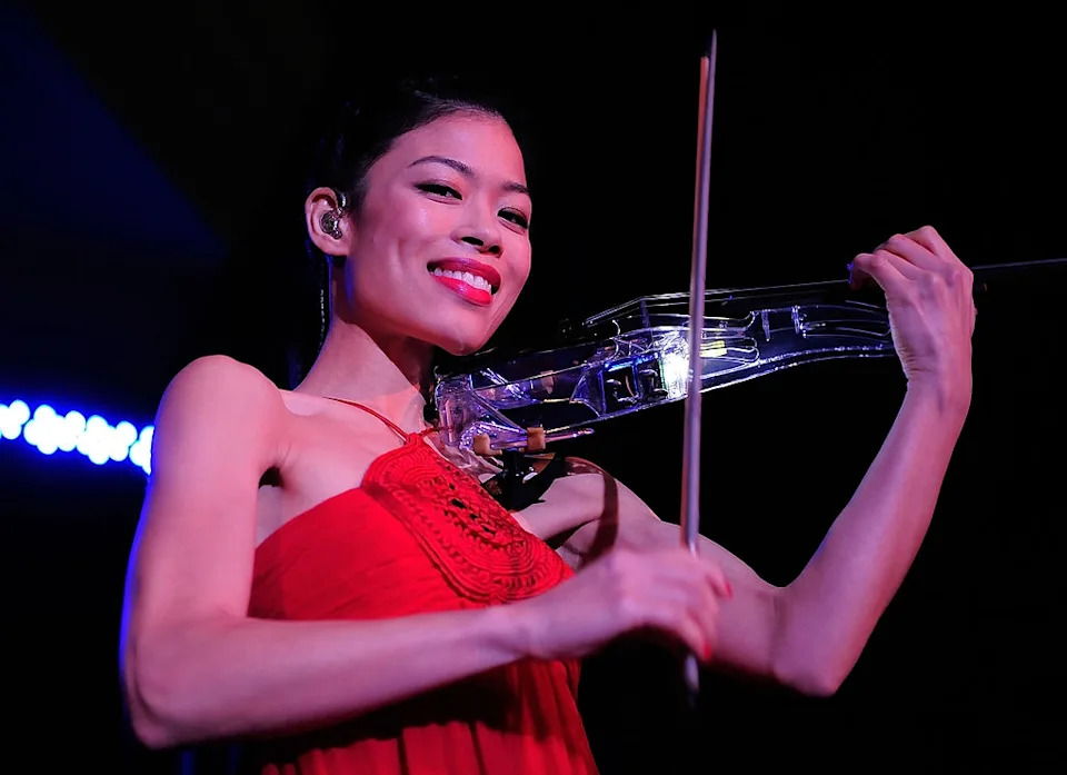A woman plays a transparent violin on stage, wearing an elegant sleeveless dress. She smiles while performing, captured in an engaging moment