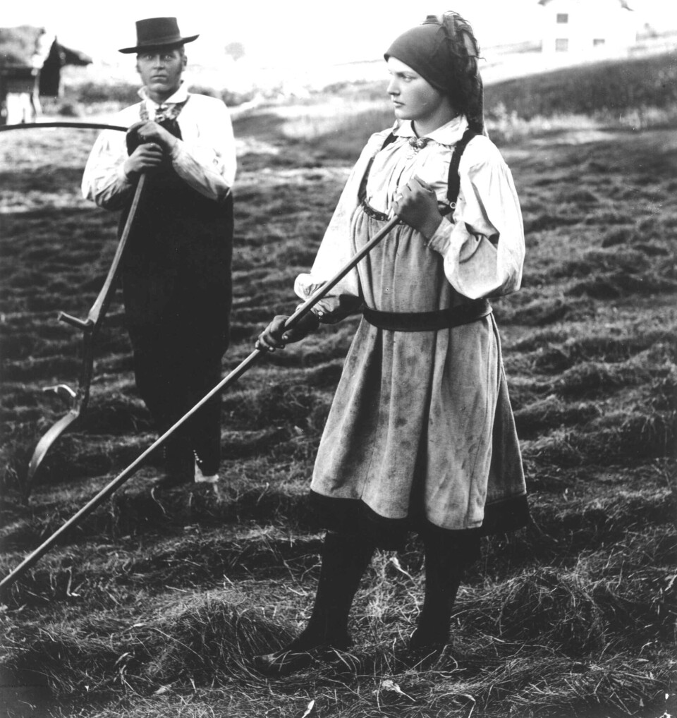 Mann og kvinne i folkedrakt med ljå og rive på jorde.