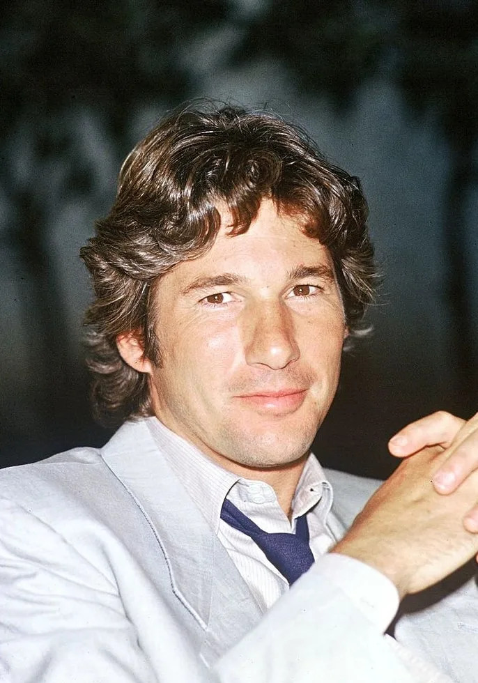 A person with curly hair in a light suit jacket poses for a portrait, hands clasped, with a neutral expression