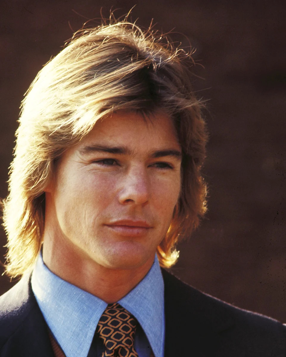 A man in a suit with long, styled hair looks ahead with a neutral expression