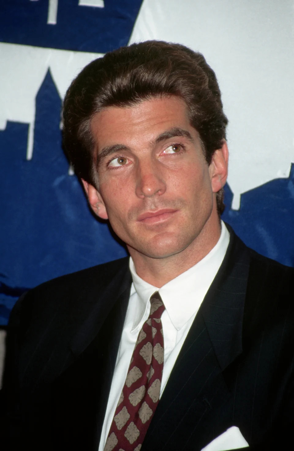 A man in a suit and tie looks to the side at a formal event, with a backdrop of a city silhouette