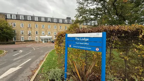 STEVE HUBBARD/BBC The Lodge in Essex is the headquarters of the Essex Partnership University NHS Foundation Trust and is shown in this picture. It is made up of light yellow bricks and is four storeys tall. It is a long building with trees to the front of it. There is a car in the immediate foreground in the car park. The window frames are white. There is an access road to the front of the picture.