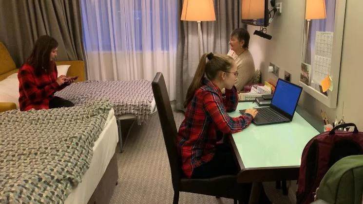 Kateryna studies in a hotel room on the only desk with her mother sat on the bed and grandmother sat in a chair in the room. 