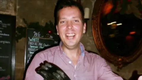 Family handout A man is looking at the camera wearing a red shirt. He is smiling. He appears to be a in a dark pub.