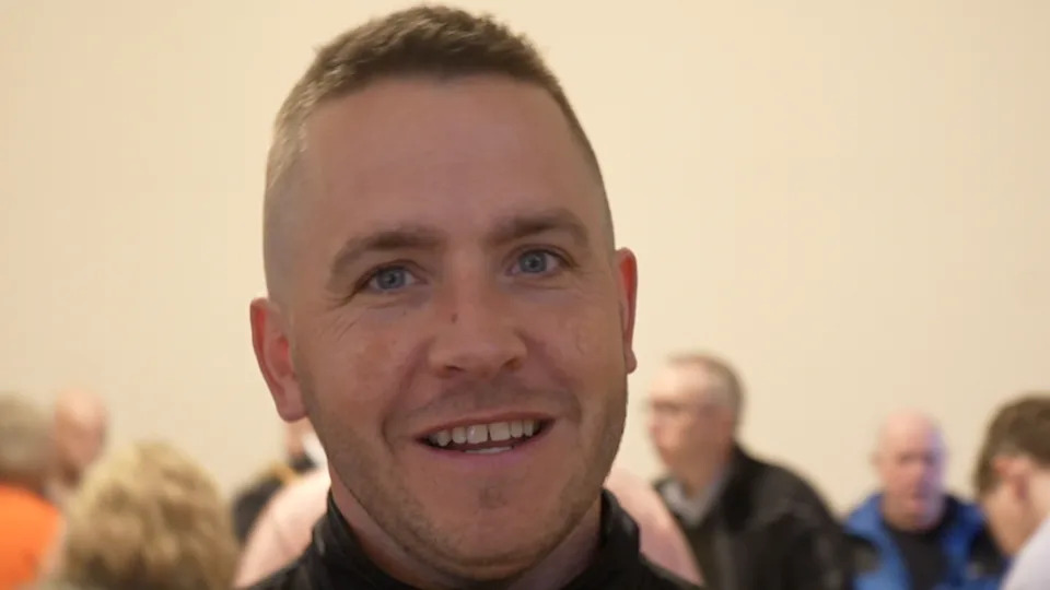 Ritchie Hawkins smiles at the camera inside a room. He has short light brown hair and some stubble. He wears a black top with a collar. 