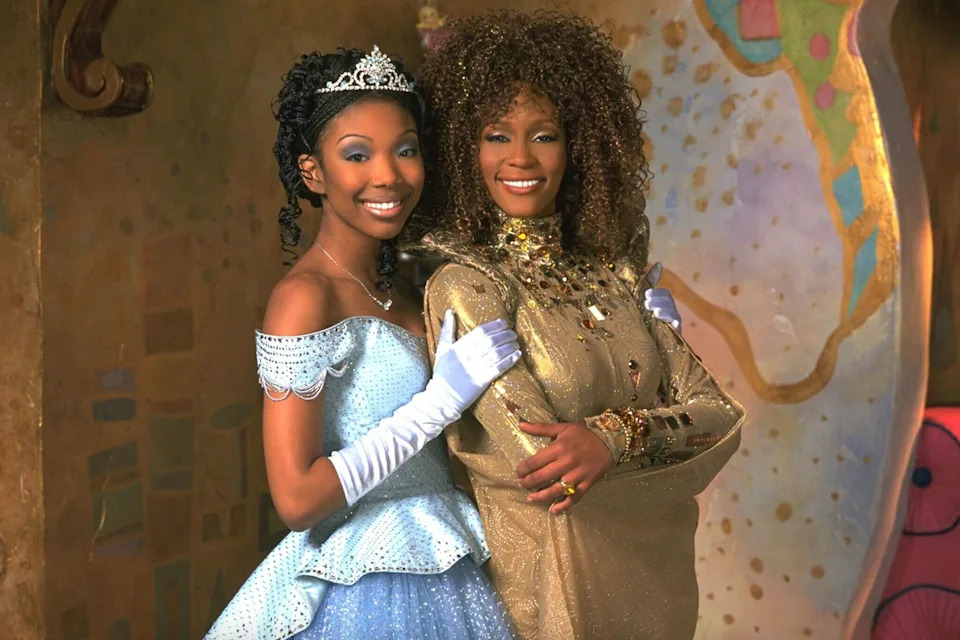 Two people in glamorous costumes; one is in a sparkling gown and tiara, the other in a gold dress with elegant jewelry, posing together