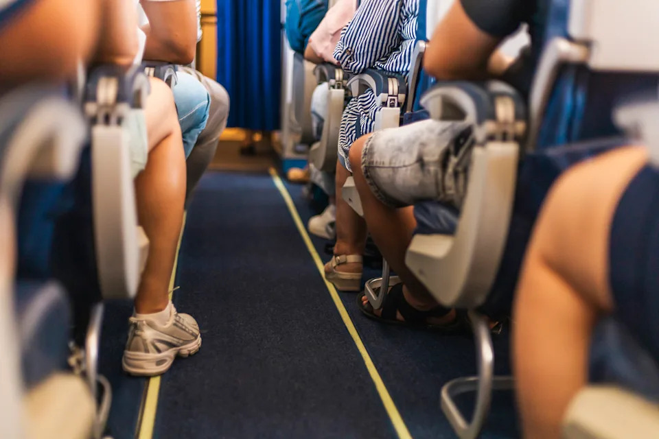 Travelers are seated in an airplane, with their legs visible from the aisle. The atmosphere indicates anticipation as the aircraft prepares for departure.
