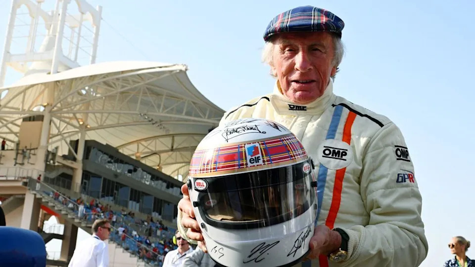 A man with short grey hair wearing a tartan cap and white racing coat and holding up a tartan and white helmet that has signatures written onto it. Stadium seating can be seen in the distance.