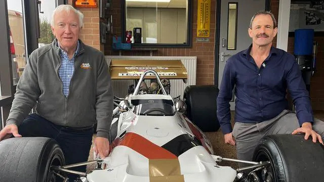 A man with short white hair wearing a grey fleece and blue shirt is leaning down with his right hand on the wheel of a white racing car. To the right is another younger man with a navy shirt, brown short hair and moustache, with his hand on the right wheel.