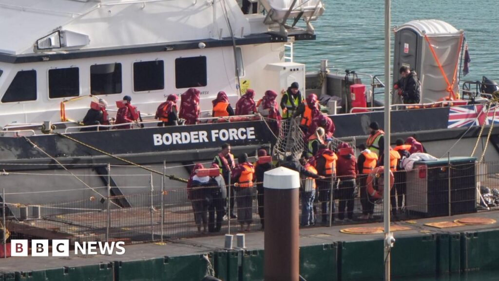 People wearing orange life jackets standing on a white boat. There are people standing on a pier.