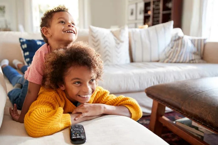 Two young children smiling and watching TV on a sofa; one is resting on the other's back. A remote control is in front of them